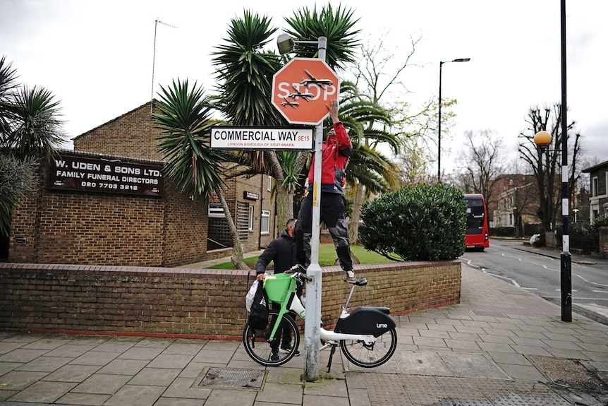 A man stands on a bike, held by another man, as he unscrews a stop sign that had been used as a canvas by Banksy