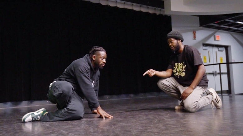 A man dressed in a black sweat shirt and black paints is crouched down on a dance studio floor. He rests his right thigh on the floor, and his left and right hands are on the floor for balance. He has tight braids in his hair. A second man, crouched beside him is pointing down towards the floor. He is wearing a black T-Shirt with yellow graphic lettering and light-coloured track pants. Both men are wearing sneakers