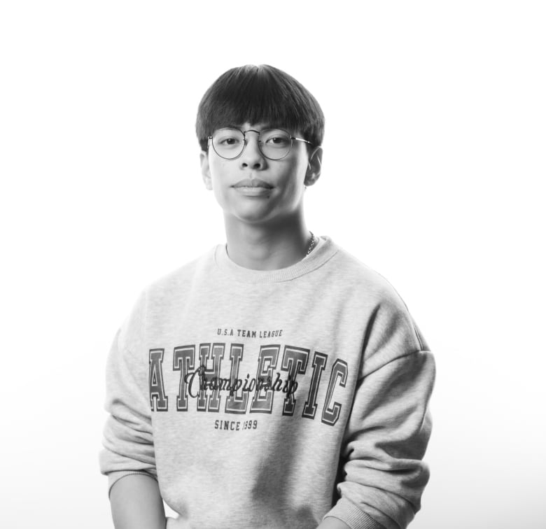Young man with short, straight hair and bangs sits on a stool. He is wearing a sweatshirt that says Athletic Championships on the front and cargo pants with a pocket on the side. He wears round metal framed glasses and has his hands clasped on his lap.