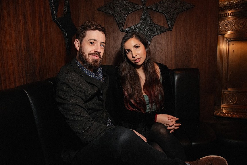 bearded man in black jacket and brunette woman pose for photo on a black leather chair