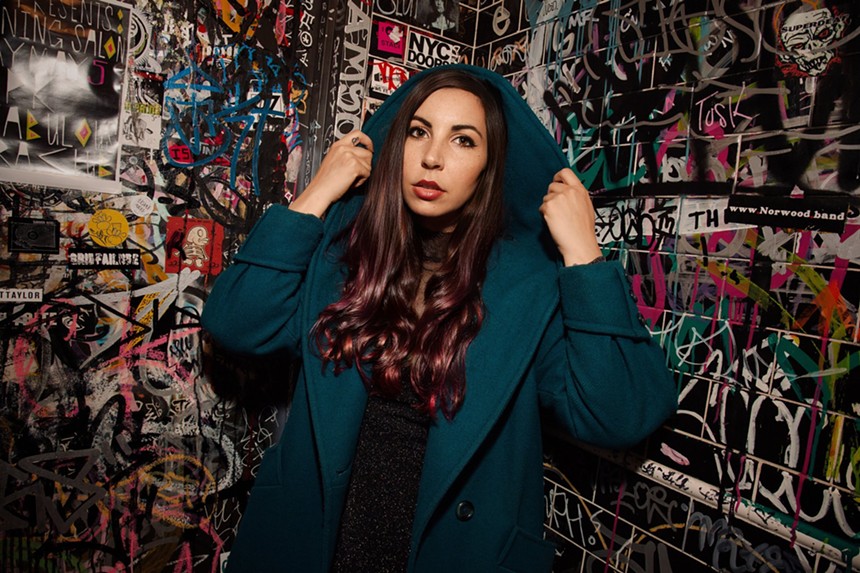 woman in blue sweater stands in front of a wall covered in graffiti.