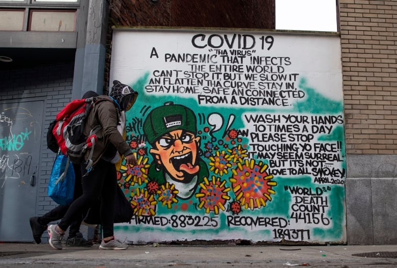 Two people walk past a mural urging health precautions to stop the spread of COVID-19.