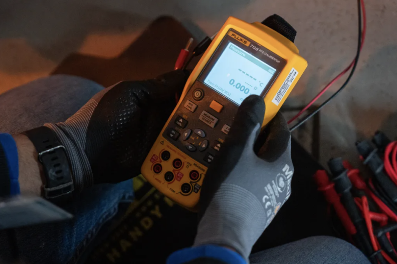 A pair of gloved hands holds a yellow module with buttons and a small lit screen that showcases a variety of data.