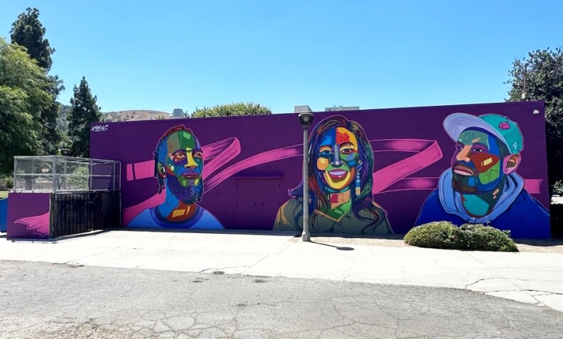 Photo of a vibrant wall, you see a purple backdrop with a streak of pink. The painted streak has 3 painted people in its forefront. One is wearing a blue tee, sporting a beard and braided hair that reaches their neck. The person is a center, who has a bright white smile, with long dark hair that reaches their shoulders, wearing a green top that isn't clearly identifiable. The one to the right of the frame wears a dark blue sweater and light blue hat, they also are sporting a goatee beard.