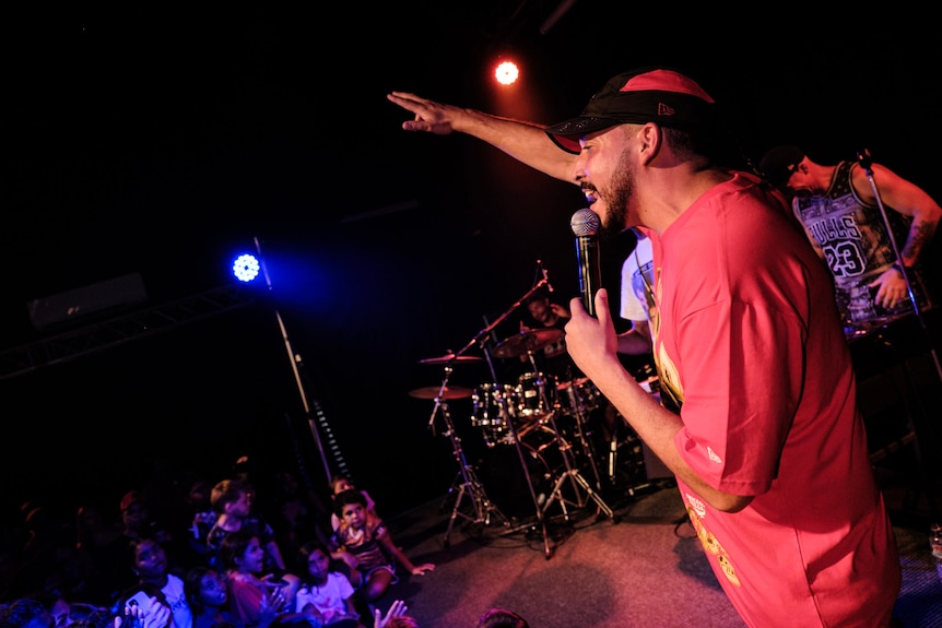 Man wearing red shirt holding microphone raps on stage with hand in the air under stage lights.