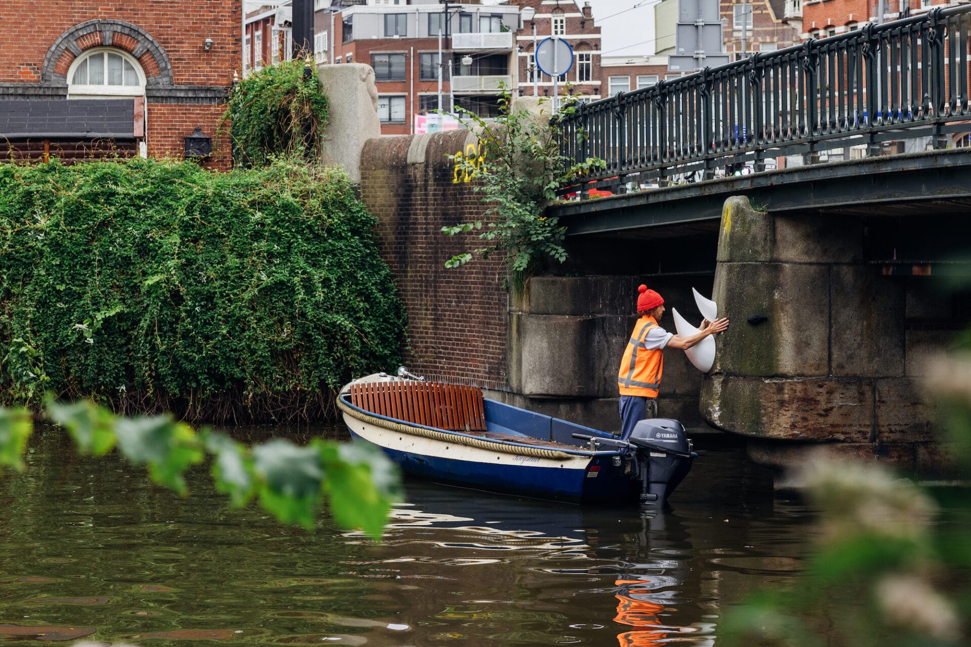 Artist Frankey stands in a boat in an Amsterdam canal and places sculptural pieces onto the side of a bridge