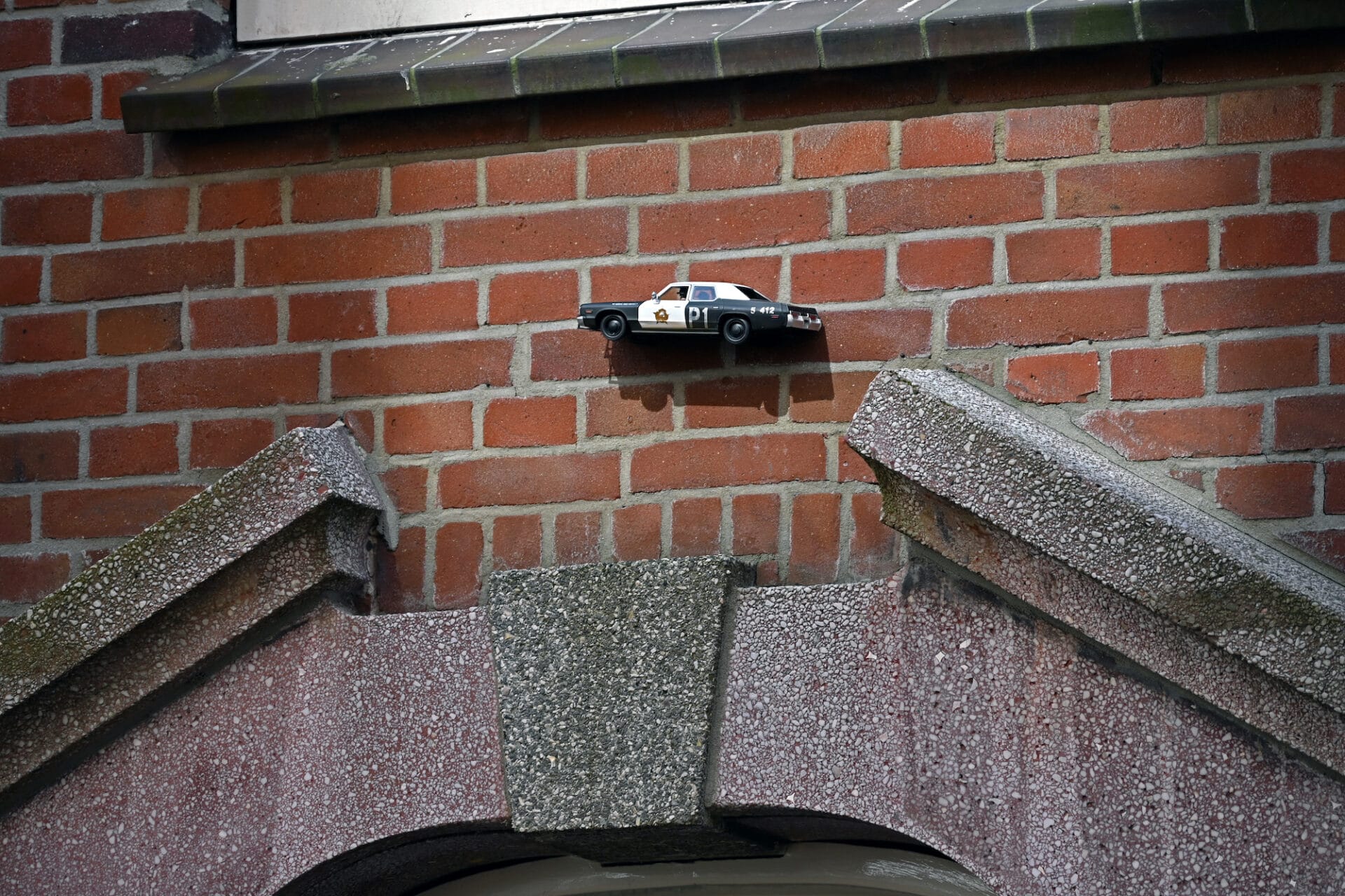 A little sculpture of a police car is stuck to a brick wall and appears to leap over a door.