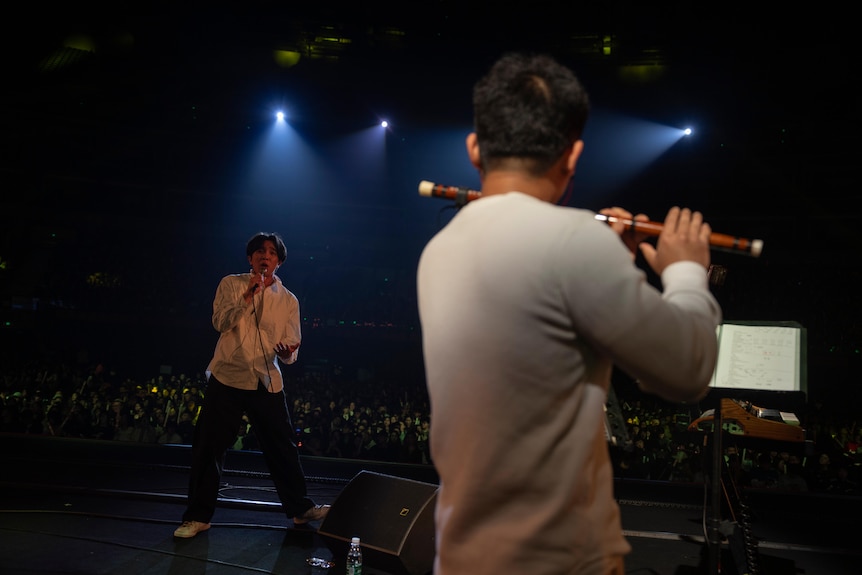 A young Chinese man raps into a microphone while another plays the flute on a stage.