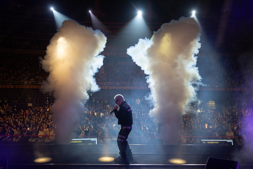 A young Chinese man with bleached blond hair sings into a microphone as smoke rises in two pillars around him on a stage.