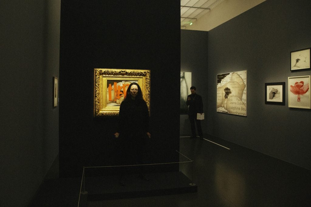 a woman stands in a gallery in front of a painting tagged with red graffiti. There are more works of art in the background.