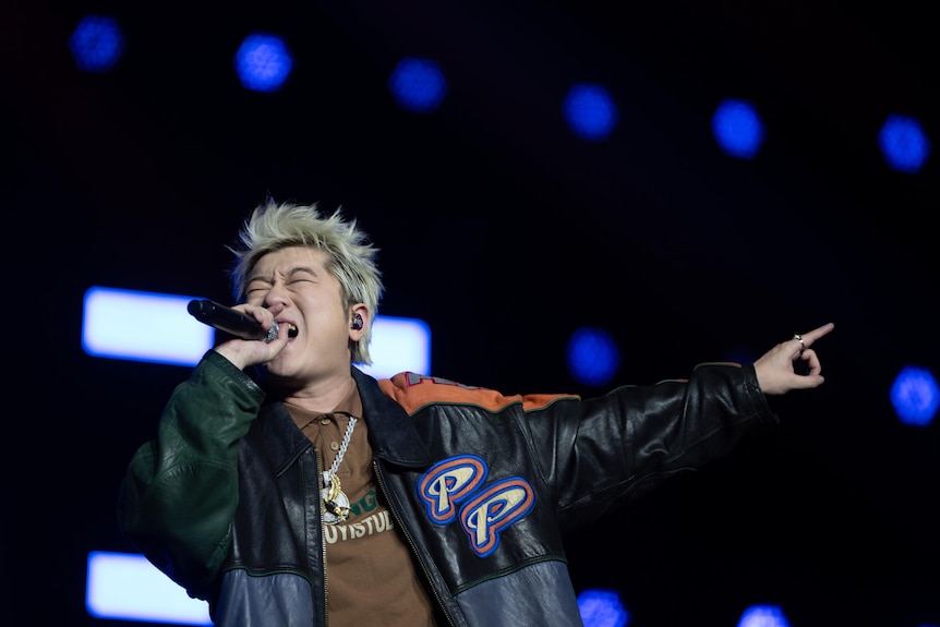 A young Chinese man with blonde hair screams into a microphone and points to the sky.