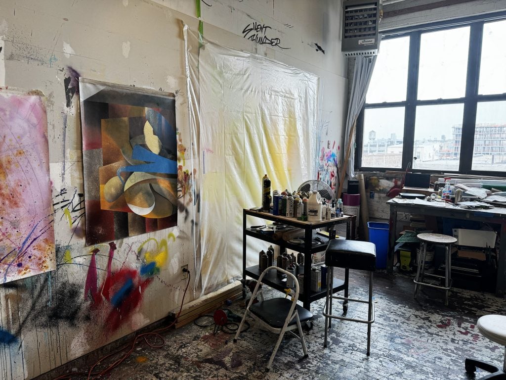 A small studio space with paintings hung on walls, a table filled with spray cans, and a floor coated in paint.