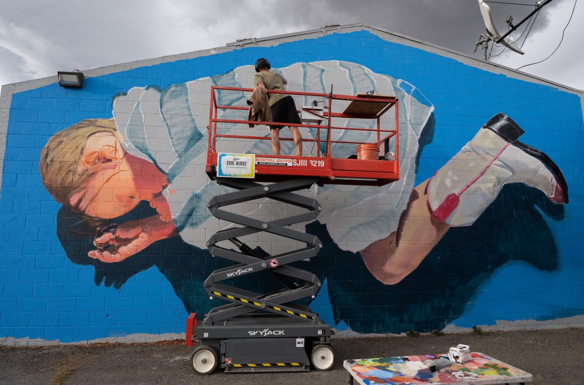 Nevada murals. Artist on cherry picker is painting a large baby in the foetal position and wearing cowboy boots on a wall.