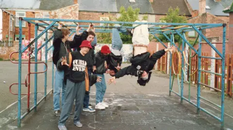 Paul Heaton Six boys playing on a metal climbing frame, which is on tarmac with a row of terraced houses in the distance