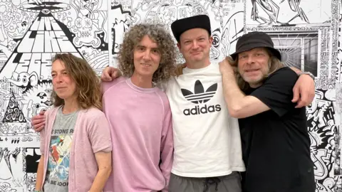 Four artists: a female with long mousey brown hair on the left, standing next to a man with curly grey hair and wearing a pink top, who is standing next to a man wearing a black baseball cap and white Adidas t-shirt, who is standing next to a slightly older man wearing a black hat and t-shirt. All four are standing in front of a graffiti montage on a wall