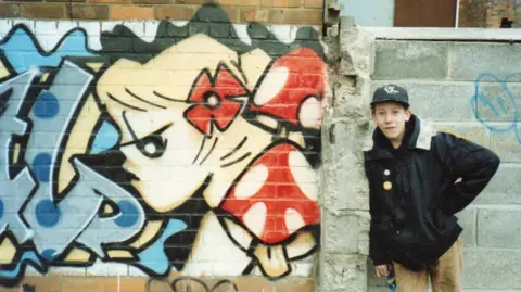 Paul Heaton Boy wearing baseball cap and black jacket is standing next to a painted wall with a cartoon depiction