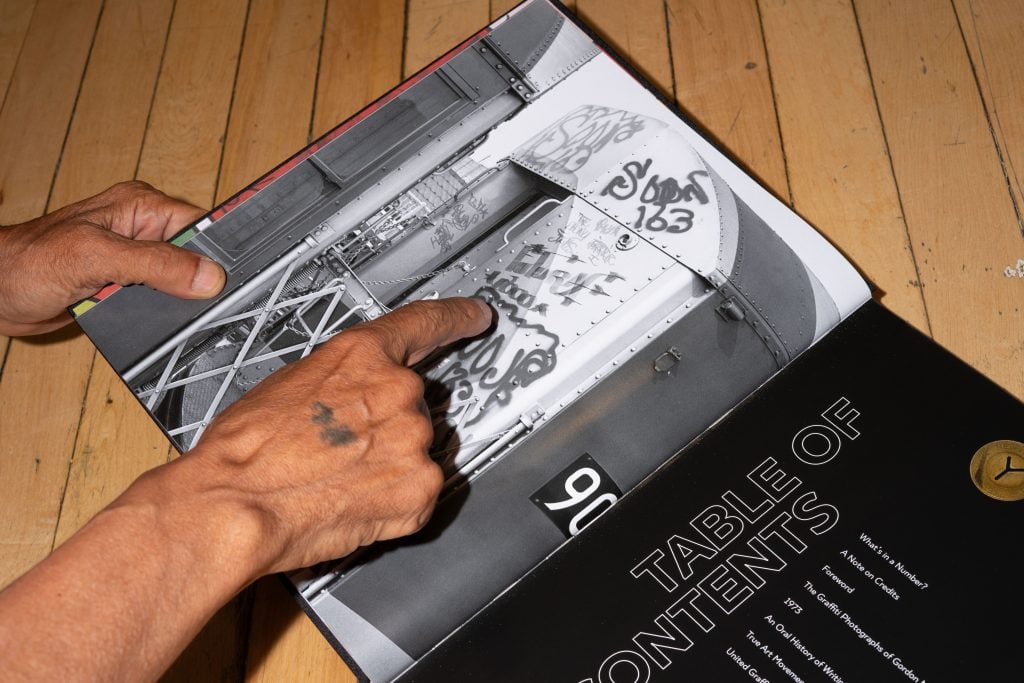 A person with a tattoo on their hand is pointing to graffiti art on a black-and-white photograph of a train car in an open book. The book's table of contents is visible on the right page, listing topics such as 