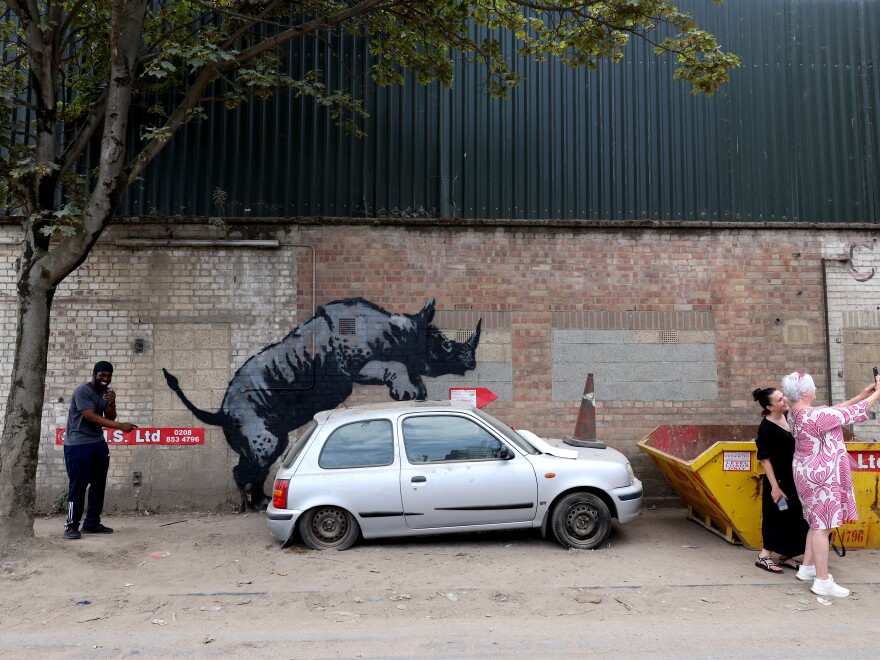 People gather around an artwork by street artist Banksy, the eighth to released in eight days, depicting a rhino mounting a car, on a wall in Charlton, London, on August 12.