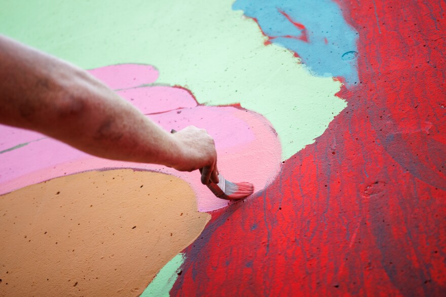 Jason Spencer, “Killer Napkins”, paints his artwork along the Mississippi River on Friday, August 30, 2024. “Just goofy with this… What if they went on vacation and they were holding a hotdog” says Spencer.