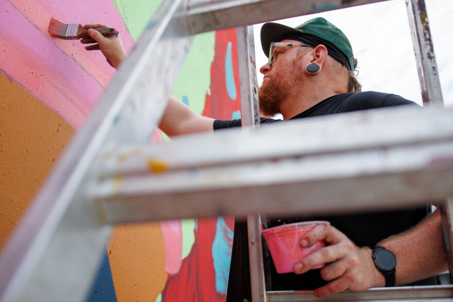 Jason Spencer, “Killer Napkins”, paints his artwork along the Mississippi River on Friday, August 30, 2024. “Just goofy with this… What if they went on vacation and they were holding a hotdog” says Spencer.