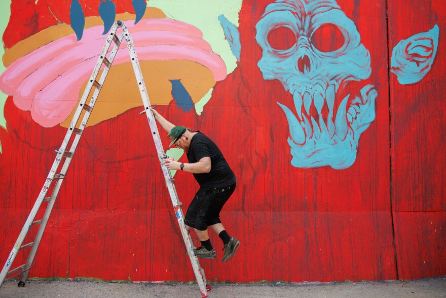 Jason Spencer, “Killer Napkins”, gets down from the ladder from painting the hotdog along the Mississippi River on Friday, August 30, 2024. “Just goofy with this… What if they went on vacation and they were holding a hotdog” says Spencer.