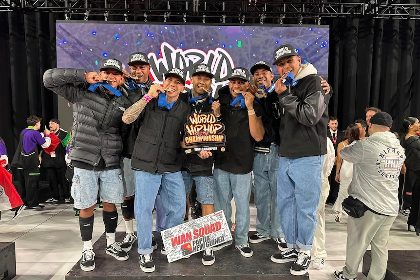 Seven men stand on the podium biting their gold medals, dressed in hip hop style clothes.