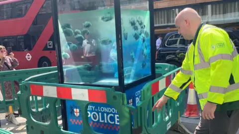 PA Media A City of London police sentry box painted in semi-opaque blue with piranhas in water, next to green, red and white barriers and a workman in yellow fluorescent clothing