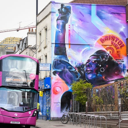 A pink bus travels past a mural of a woman holding an arm aloft in triumph, with a disc behind her like a halo saying Bristol Rise Up