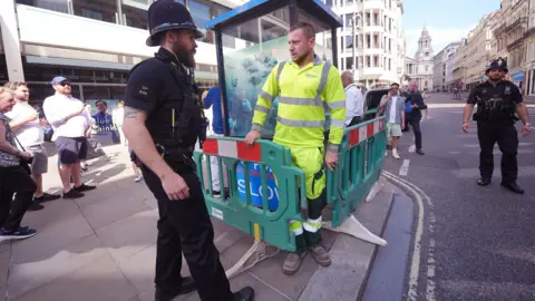 PA Media Contractor wearing high vis top and trousers erecting barrier round box while a police officer stands by. Passers by are watching the scene from the pavement.