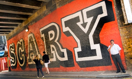 A man smoking a cigarette and a couple are dwarfed by the word SCARY spray painted under a bridge