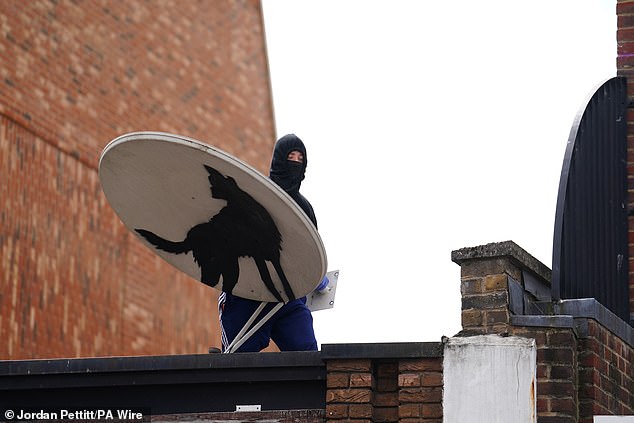 A hooded man with a mask holding the satellite dish while stood on top of the building