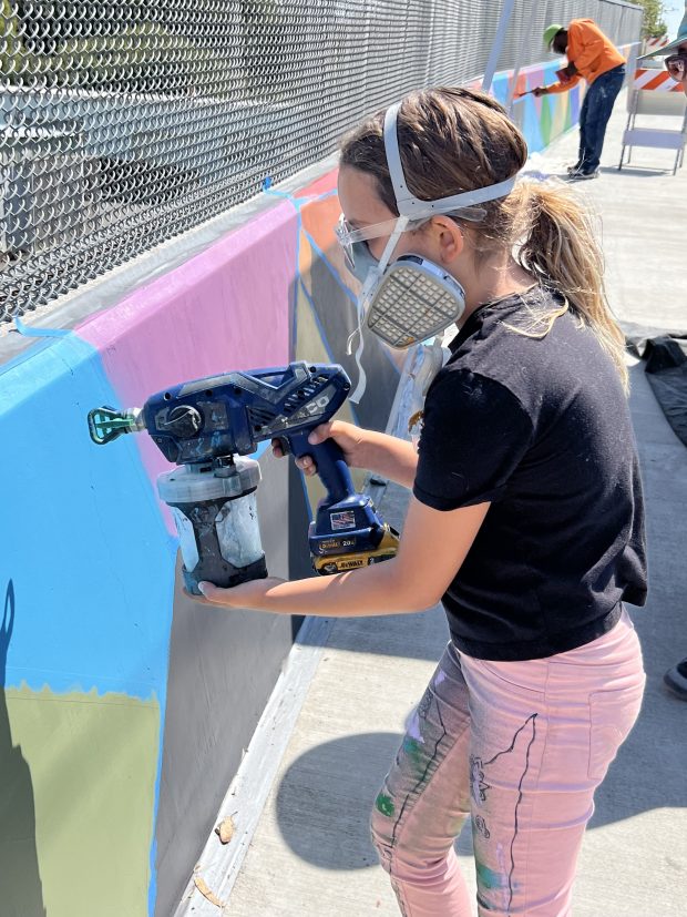 Edin Goulart, 8, wears protective gear as she demonstrates her father's new paint sprayer on the Hawthorne Street mural. She worked on the mural with her dad, Ben Goulart, as part of the Eureka Street Art Festival in 2023. (Heather Shelton/The Times-Standard file)