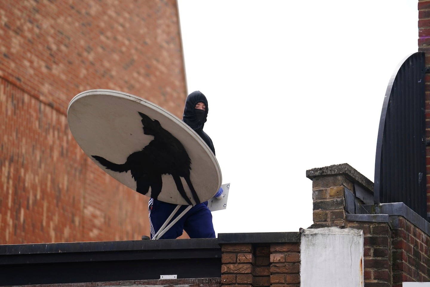 A covered-face person removes a new artwork by Banksy, depicting a howling wolf painted on a satellite dish that was placed on a shop roof in Peckham, south London, Thursday Aug. 8, 2024.