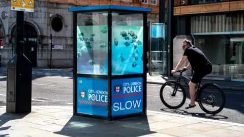 Bansky Banksy work of fish shoal, possibly piranhas, painted onto a glass police sentry box. The view is taken from the pavement and on the right, a cyclist rides by.