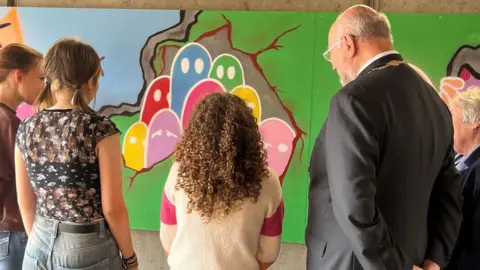 City of Ely Council The back view of three teenagers and a man wearing a mayoral chain over his grey suit and a woman with grey hair looking at part of the finished mural