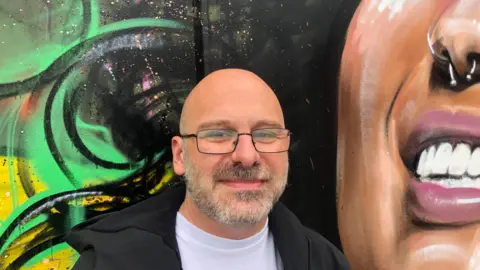 John Devine/BBC Dave Farrington, 41wears a brown/grey beard and moustache with light framed glasses, a black jacket over a white T-shirt. He is standing in front of part of the mural featuring the smiling woman's lower face.