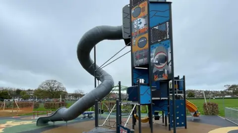 Torridge District Council Tall play tower with different activities. There is a grey slide with a twist and grass in the background.