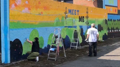 LDRS Residents taking off paint - a woman in the foreground on her knees scrubbing at a wall with two stepladders near the mural which says meet me.