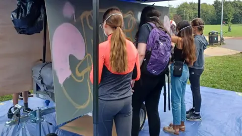 City of Ely Council The back views of four teenagers, all with long hair, working on an artwork on a large board in a field, while standing on a blue plastic dustsheet