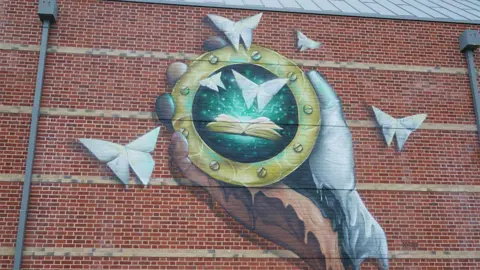 Wild Walls Festival/Joe Barton A picture shows the large scale mural created by artist Nathan Murdoch on the Suffolk Archives building The Hold. It shows a hand holding a portal suggesting the ability the time travel while origami butterflies made with folded archive records fly out of the portal.