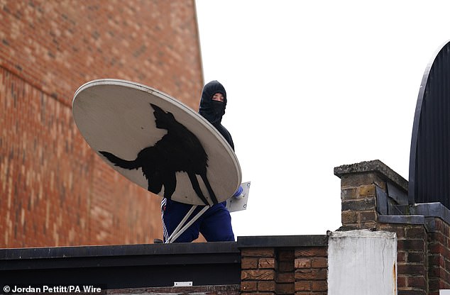 A hooded man with a mask is seen holding a satellite dish with Banksy's wolf mural on it before the work disappeared