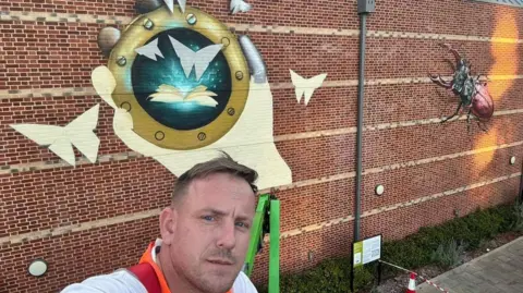 Wild Walls Festival/Nathan Murdoch Nathan Murdoch in a selfie-style picture in front of his large scale mural on the back of The Hold archives building in Ipswich. He is pictured looking at the camera while wearing an orange high-viz jacket.