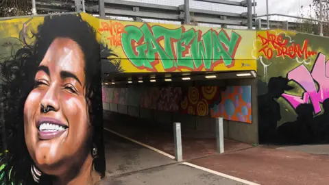 John Devine/BBC Colourful mural at the entrance to an underpass, a smiling woman has her eyes closed and appears to be in full sunshine on the left. 