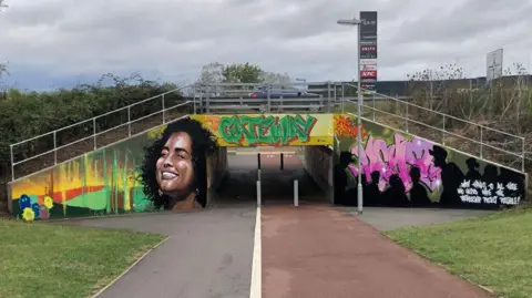 John Devine/BBC A colourful mural on the end of an underpass, showing a smiling woman on the right with her face raised, Gateway written above its entry in green letters and figures silhouetted in black against pink letters on the right, beneath the A10 with a car midway across the bridge