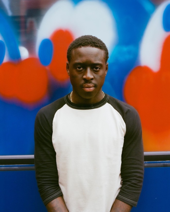 A young man wearing a white and black T-shirt stands in front of a blue bus whose windows are spray-painted in blue, white and red