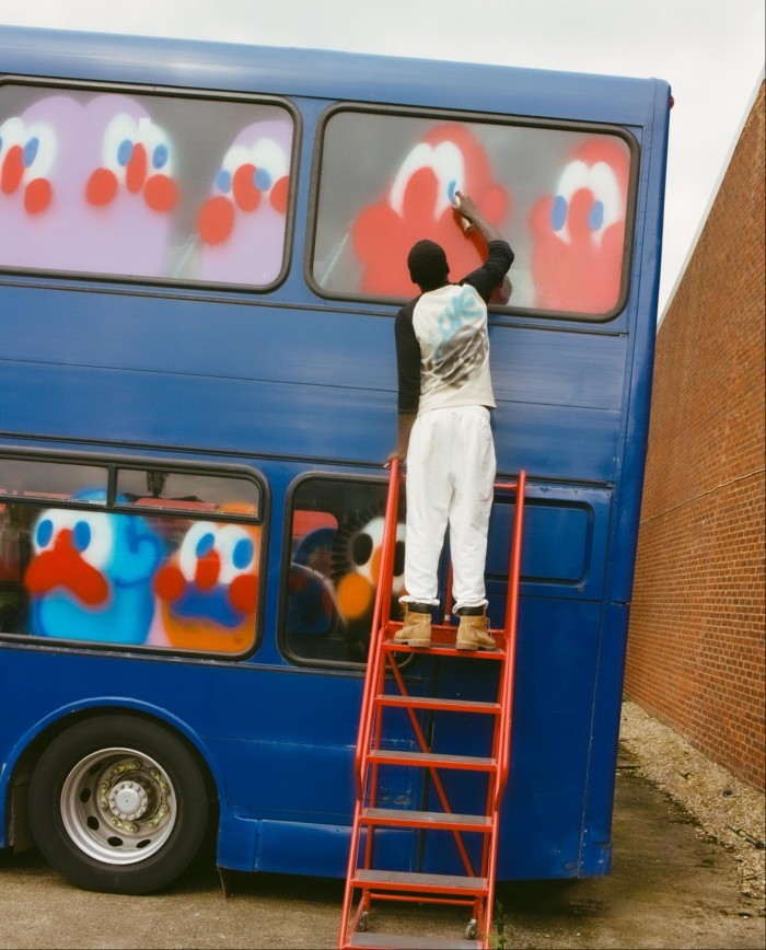 A young man wearing a white and black T-shirt, white jogging bottoms and tan-coloured work boots stands at the top of a red step ladder in front of a blue double-decker bus adding blue eyes onto faces spray-painted on the windows