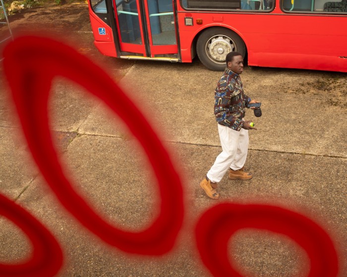 A young man wearing a brown patterned fleece top, white jogging bottoms and tan-coloured work boots is seen through a graffiti-ed window. He stands next to a red bus holding a can of spray paint and a mobile phone