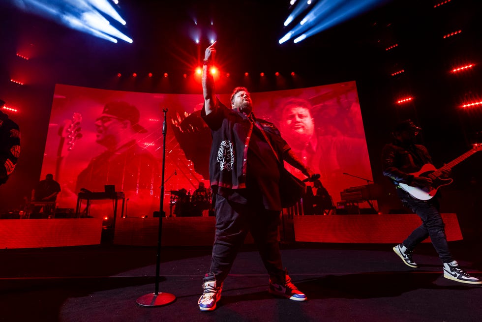 jelly roll stands on a stage with one hand pointing up in the air and another holding a microphone out at his side, he wears a black button up over a black shirt, dark pants, and sneakers, behind him is a large video screen and musicians with instruments