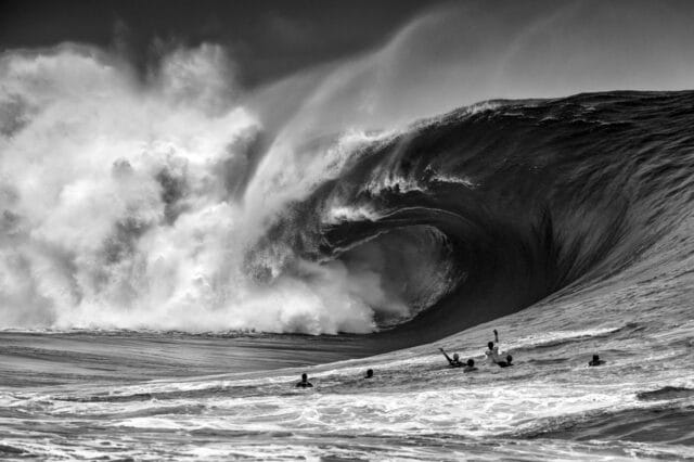 in a black and white photo, a large barreling wave begins to crash down on itself as a handful of individuals in the water nearby watch