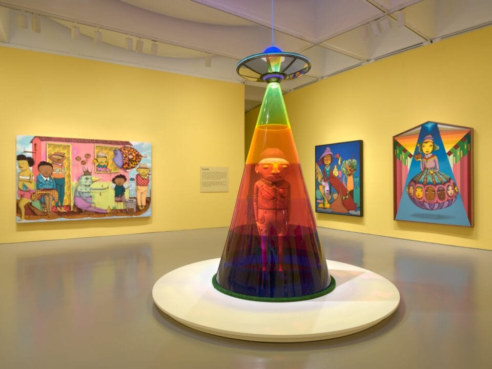 an installation view of a figurative sculpture encased in a rainbow colored beam from a flying saucer. three colorful paintings with figures line the walls behind